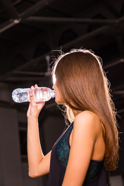 Jovem aptidão bebendo água da garrafa. Mulher jovem e musculosa no ginásio, fazendo uma pausa do treino.