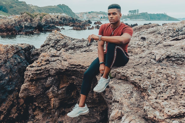 Jovem apto segurando seus óculos de sol nas mãos enquanto está sentado na rocha perto do mar calmo