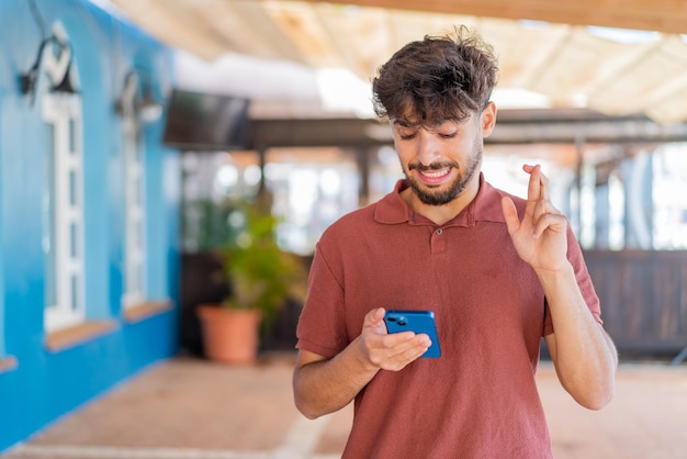Jovem árabe bonito ao ar livre usando telefone celular com os dedos cruzados