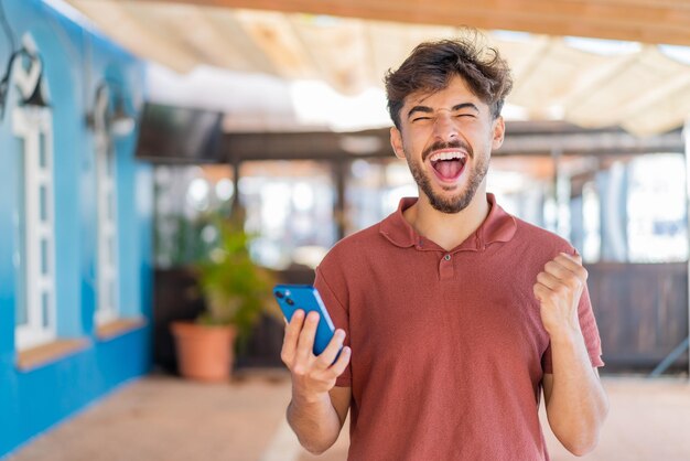 Foto jovem árabe bonito ao ar livre usando telefone celular e fazendo um gesto de vitória