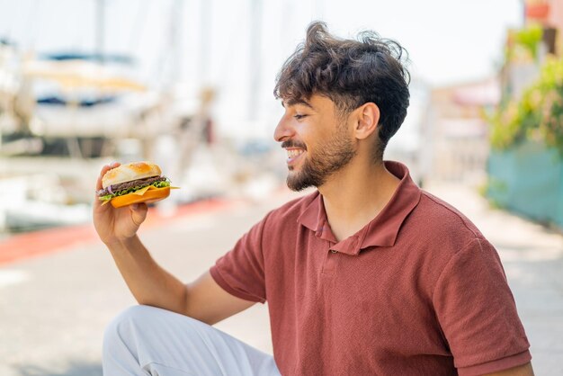 Foto jovem árabe bonito segurando um hambúrguer ao ar livre com expressão feliz