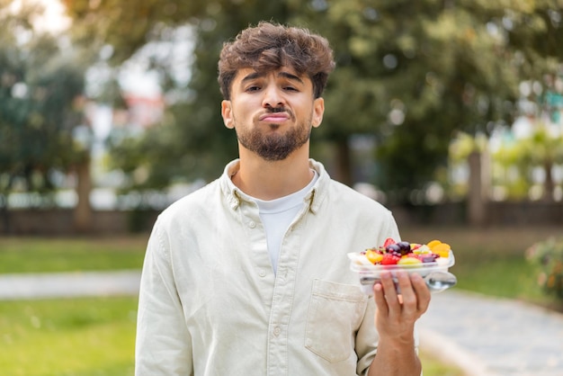 Jovem árabe bonito segurando uma tigela de frutas ao ar livre com expressão triste