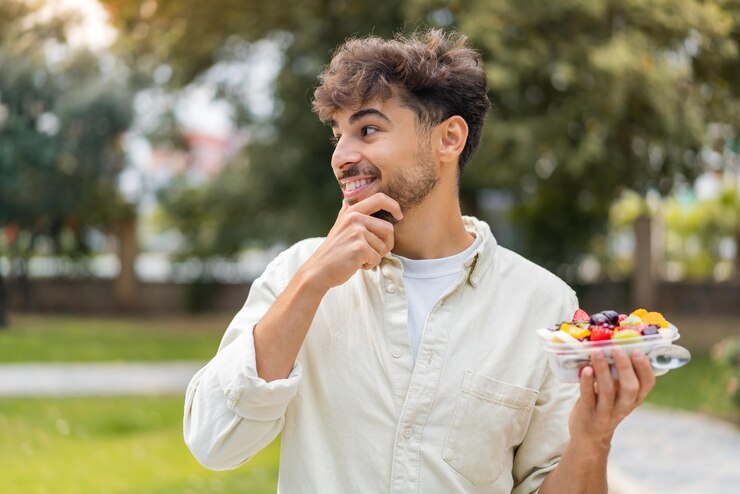 Foto jovem árabe bonito segurando uma tigela de frutas ao ar livre pensando em uma ideia e olhando de lado
