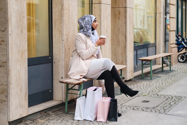Jovem árabe elegante em hijab bebendo café no banco na rua da cidade depois de fazer compras