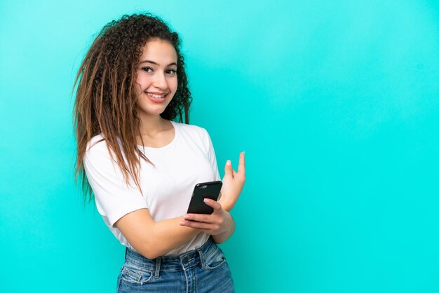 Jovem árabe isolada em fundo azul usando telefone celular e apontando para trás