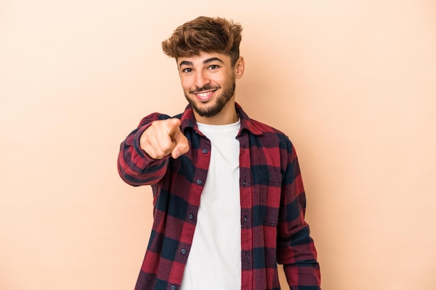 Jovem árabe isolado em sorrisos alegres de fundo bege, apontando para a frente.