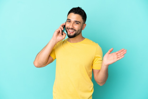 Jovem árabe isolado em um fundo azul, conversando com alguém ao telefone celular.