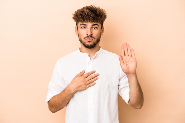 Jovem árabe isolado em um fundo bege, fazendo um juramento, colocando a mão no peito.