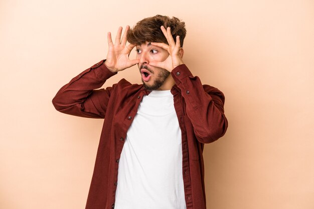 Jovem árabe isolado em um fundo bege, mantendo os olhos abertos para encontrar uma oportunidade de sucesso.