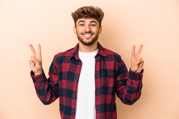 Jovem árabe isolado em um fundo bege, mostrando o sinal da vitória e sorrindo amplamente.