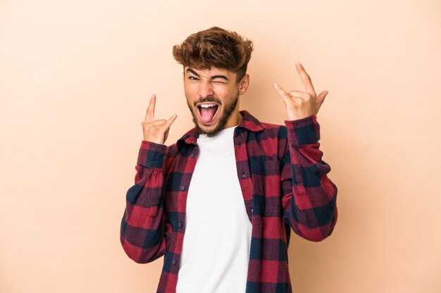 Jovem árabe isolado em um fundo bege, mostrando um gesto de chifres como um conceito de revolução.