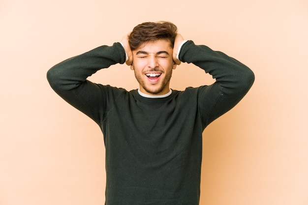 Jovem árabe isolado em um fundo bege ri com alegria, mantendo as mãos na cabeça. Conceito de felicidade.