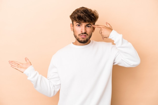 Jovem árabe isolado em um fundo bege, segurando e mostrando um produto por lado.