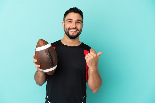 Jovem árabe jogando rugby isolado em um fundo azul apontando para o lado para apresentar um produto