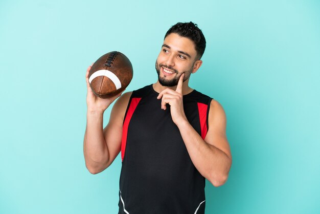 Jovem árabe jogando rugby isolado em um fundo azul tendo uma ideia enquanto olha para cima