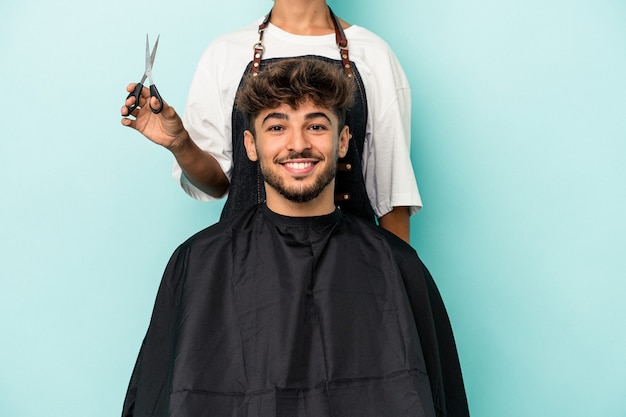 Jovem árabe pronto para cortar o cabelo isolado em um fundo azul feliz, sorridente e alegre.