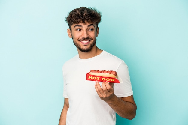 Jovem árabe segurando um cachorro-quente isolado em um fundo azul parece de lado sorrindo, alegre e agradável.