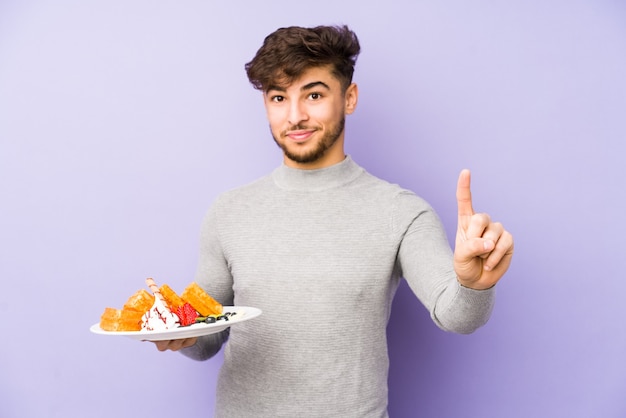 Jovem árabe segurando um waffle isolado mostrando o número um com o dedo.