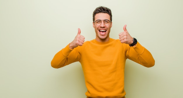 Jovem árabe sorrindo amplamente olhando feliz, positivo, confiante e bem sucedido, com os dois polegares contra a parede plana