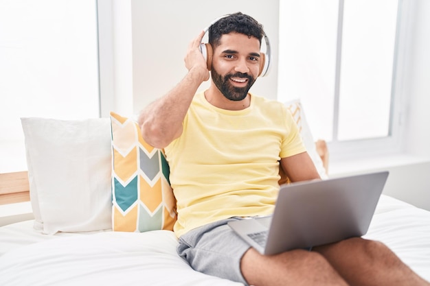 Jovem árabe usando laptop e fones de ouvido sentado na cama no quarto