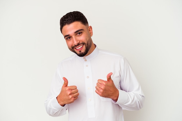 Jovem árabe vestindo roupas típicas árabes, isoladas na parede branca, levantando os dois polegares, sorrindo e confiante.