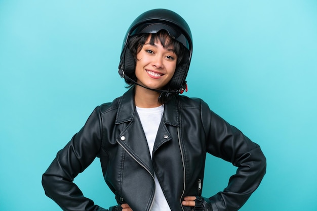 Jovem argentina com um capacete de moto isolado em fundo azul posando com braços no quadril e sorrindo