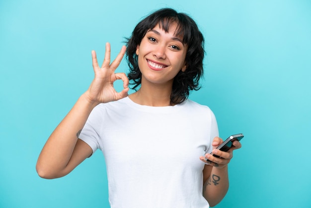 Jovem argentina isolada em fundo azul usando telefone celular e fazendo sinal de OK