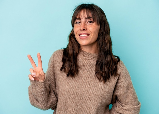 Jovem argentina isolada em um fundo azul, mostrando sinal de vitória e sorrindo amplamente.