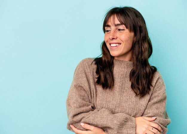 Jovem argentina isolada em um fundo azul, sorrindo confiante com os braços cruzados.