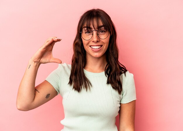 Jovem argentina isolada em um fundo rosa segurando algo pequeno com os indicadores, sorrindo e confiante.