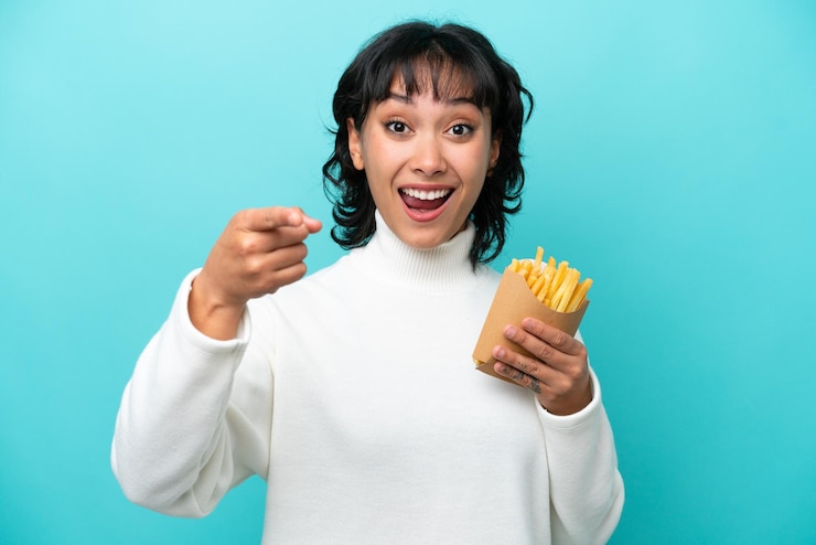 Foto jovem argentina segurando batatas fritas isoladas em fundo azul surpresa e apontando para a frente