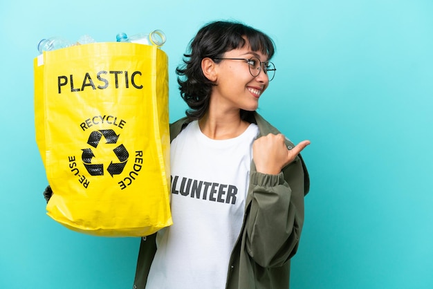 Jovem argentina segurando um saco cheio de garrafas plásticas para reciclar isolado em fundo azul apontando para o lado para apresentar um produto