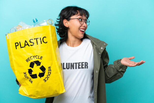 Jovem argentina segurando um saco cheio de garrafas plásticas para reciclar isolado em fundo azul com expressão de surpresa enquanto olha de lado