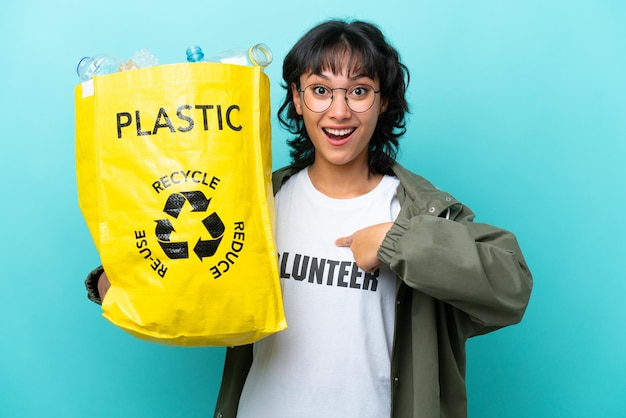 Jovem argentina segurando um saco cheio de garrafas plásticas para reciclar isolado em fundo azul com expressão facial de surpresa