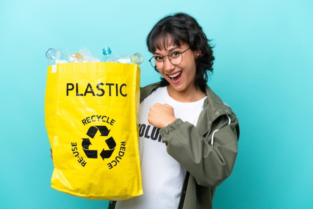 Jovem argentina segurando um saco cheio de garrafas plásticas para reciclar isolado em fundo azul comemorando uma vitória