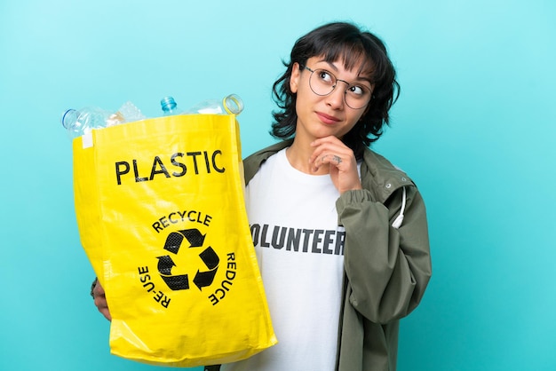 Jovem argentina segurando um saco cheio de garrafas plásticas para reciclar isolado em fundo azul olhando para cima enquanto sorria