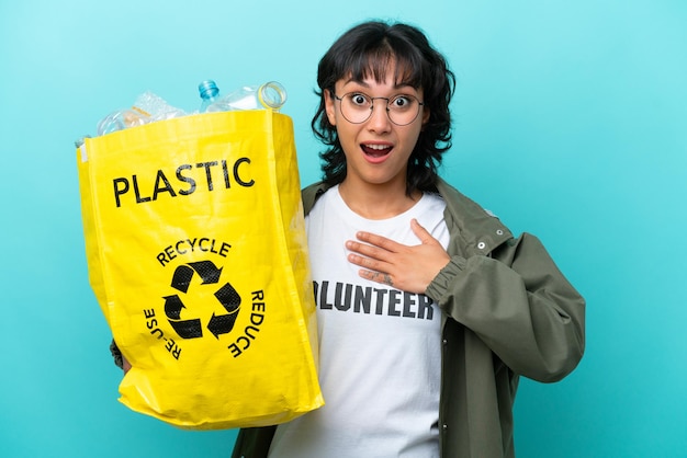 Jovem argentina segurando um saco cheio de garrafas plásticas para reciclar isolado em fundo azul surpreso e chocado ao olhar para a direita