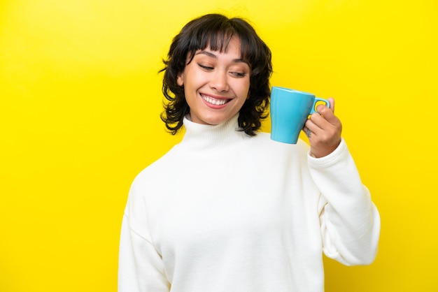 Jovem argentina segurando uma xícara de café isolada em fundo amarelo com expressão feliz