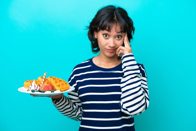 Jovem argentina segurando waffles isolados em fundo azul, pensando em uma ideia