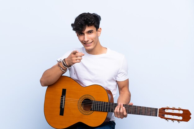 Jovem argentino com guitarra sobre parede azul isolada aponta o dedo para você com uma expressão confiante