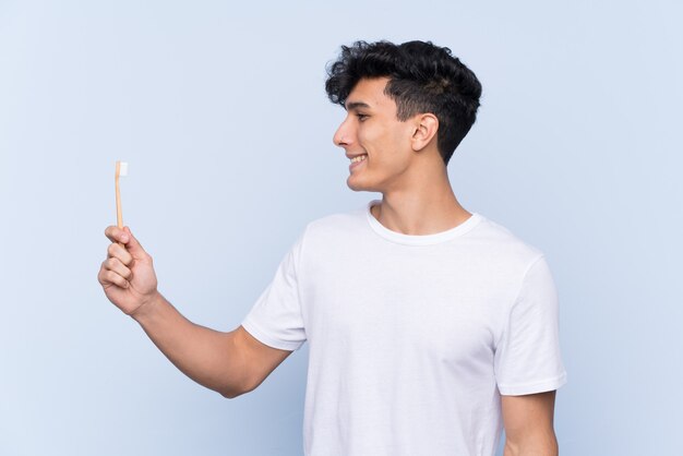 Jovem argentino escovando os dentes sobre parede azul isolada com expressão feliz