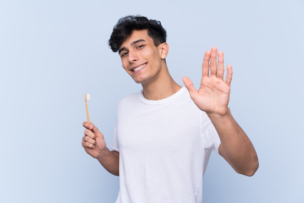 Jovem argentino escovando os dentes sobre parede azul isolada, saudando com a mão com expressão feliz