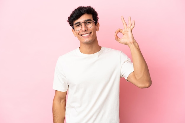 Jovem argentino isolado em fundo rosa, mostrando sinal de ok com os dedos