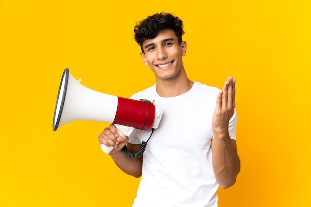 Jovem argentino isolado em um fundo amarelo segurando um megafone e convidando para vir com a mão