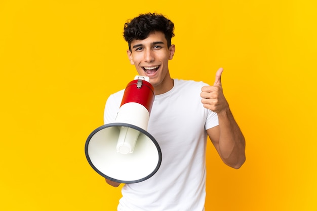 Jovem argentino isolado na parede amarela gritando em um megafone para anunciar algo e com o polegar para cima