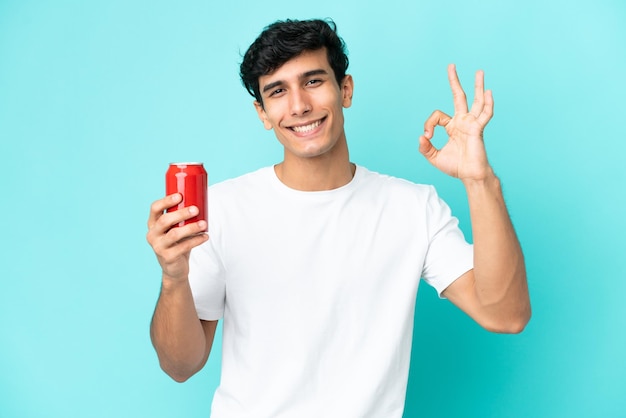 Jovem argentino segurando um refresco isolado em fundo azul, mostrando sinal de ok com os dedos