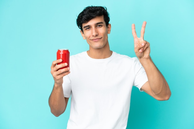 Jovem argentino segurando um refresco isolado em fundo azul sorrindo e mostrando sinal de vitória