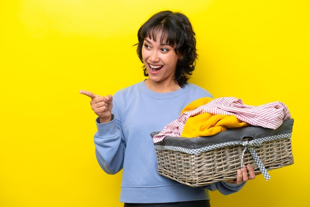 Jovem argentino segurando uma cesta de roupas isolada em fundo amarelo, apontando o dedo para o lado e apresentando um produto