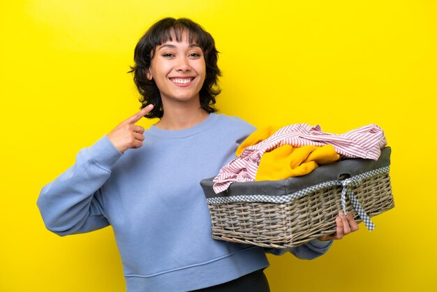 Jovem argentino segurando uma cesta de roupas isolada em fundo amarelo dando um polegar para cima gesto