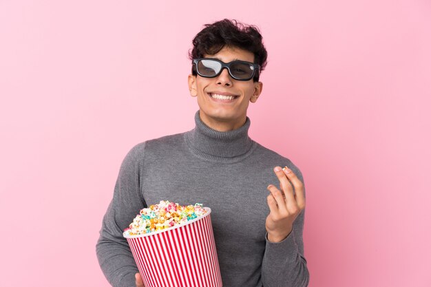 Jovem argentino sobre parede branca isolada com óculos 3d e segurando um grande balde de pipocas